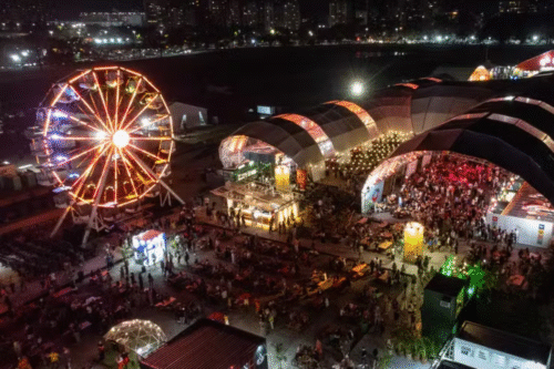 Imagem de uma feira ou parque de diversões à noite, com roda gigante iluminada e várias tendas com shows e atrações, muita gente curtindo na cidade.