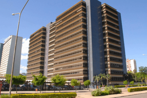 Imagem de um edifício alto de arquitetura moderna com várias janelas e uma fachada de cor escura, localizado na cidade, com céu clara ao fundo.