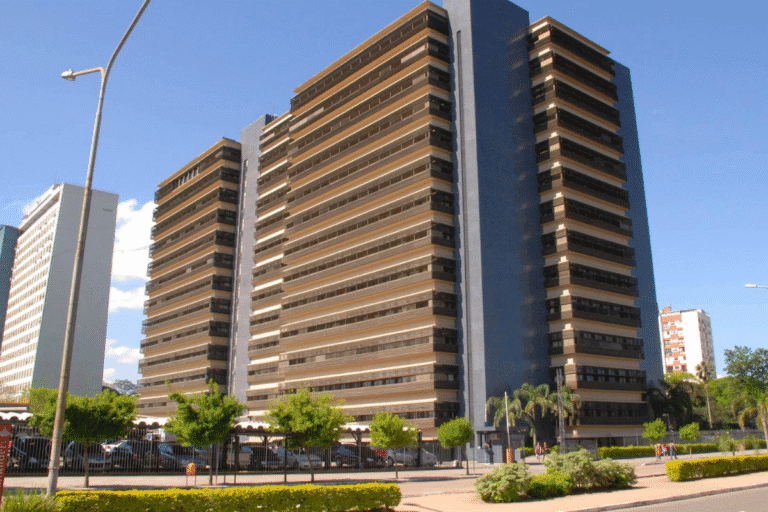 Imagem de um edifício alto de arquitetura moderna com várias janelas e uma fachada de cor escura, localizado na cidade, com céu clara ao fundo.