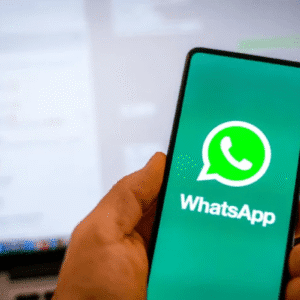 Pessoa segurando um smartphone exibindo o logo do WhatsApp com uma tela de computador ao fundo.
