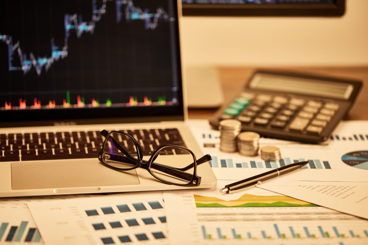 Mesa com notebook exibindo gráficos, óculos, calculadora, moedas e papéis com dados financeiros, representando erros mais comuns de investidores iniciantes