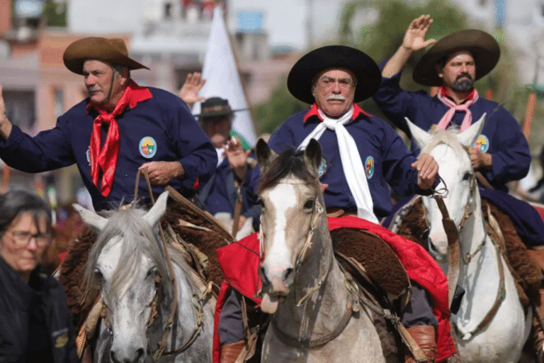 Homens vestindo roupas tradicionais de cavaleiros, montados em cavalos brancos, participando de uma celebração cultural com bandeiras ao fundo para o Dia do Gaúcho em 20 de setembro