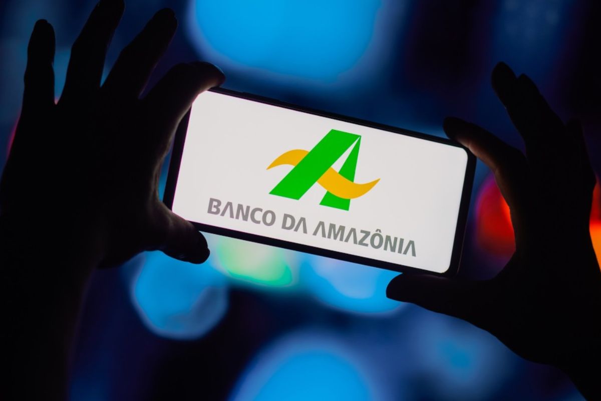 Pessoa segurando smartphone exibindo o logo do Banco da Amazônia, com fundo escuro e luzes coloridas, destaca ação de usar banco digital na região amazônica.