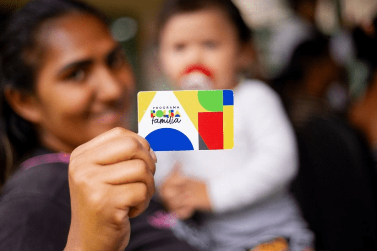 Pessoa segurando cartão do programa 'Famiília Família' em evento, com outras pessoas ao fundo, em um ambiente social e alegre.