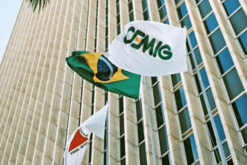 Bandeiras do Brasil e de uma organização com a palavra 'GEMIC' balançando ao vento em frente a um edifício moderno de vidro e concreto.