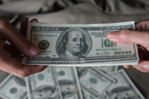 Pessoa trocando uma nota de cem dólares, destaque para a nota com o rosto de Benjamin Franklin, simbolizando dinheiro e valor econômico.