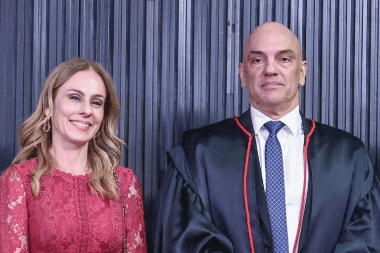 Viviane de Moraes com um vestido de renda vermelha sorri ao lado de Alexandre de Moraes com um robe preto formal com detalhes vermelhos. O fundo apresenta painéis escuros verticais, criando um ambiente formal.