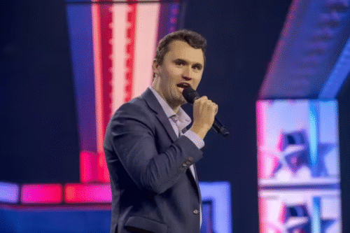 Charlie Kirk sorrindo enquanto segura um microfone, em um palco com iluminação colorida. O cenário apresenta elementos de palco em tons de rosa e azul, criando um ambiente vibrante e envolvente.