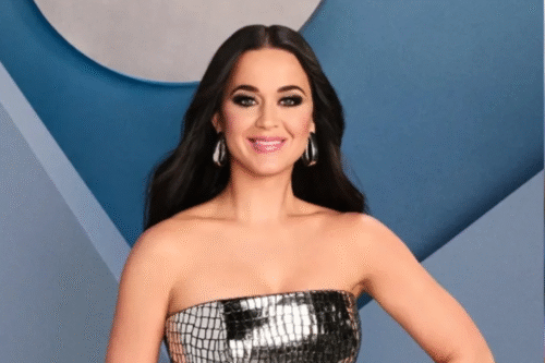 Katy Perry, sorrindo, usando vestido prata brilhante sem alças, com fundo geométrico azul e cinza, em ambiente de evento de moda.
