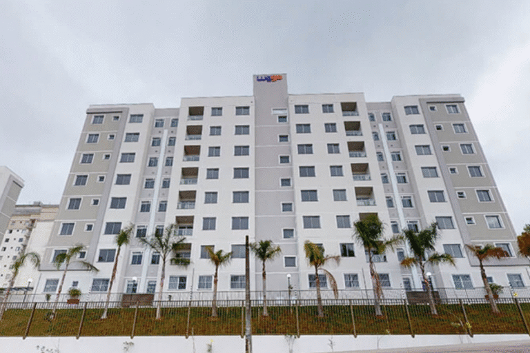 Imagem de um edifício residencial moderno com várias janelas, localizado em uma área com árvores palmadas, sob céu nublado.