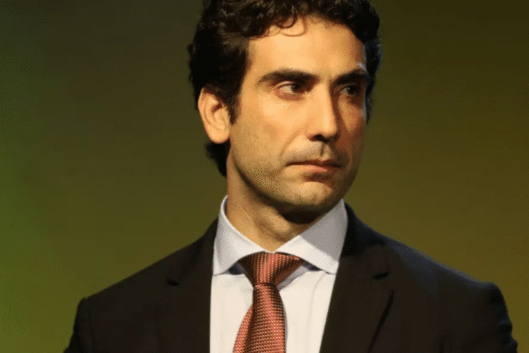 Galipolo com cabelo escuro, vestindo terno preto e gravata vermelha, olhando pensativo contra fundo amarelo.