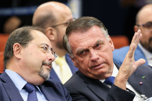 Imagem de Jair Bolsonaro em um encontro político discutindo com colegas ao seu lado, todos em traje formal e com expressão séria, ambiente de reunião pública.