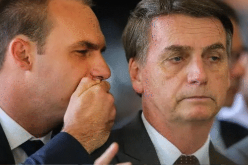 Imagem de Jair Bolsonaro e Flávio Bolsonaro em uma reunião importante, uma delas sussurrando ao ouvido da outra, dando a entender um momento de confidência ou estratégia.