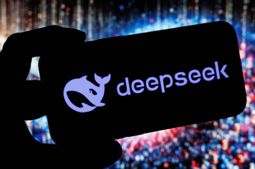 Imagem de uma tela de celular com o logo do DeepSeek iluminado, sendo segurada por uma mão, com fundo de luzes difusas em tons de azul e vermelho.