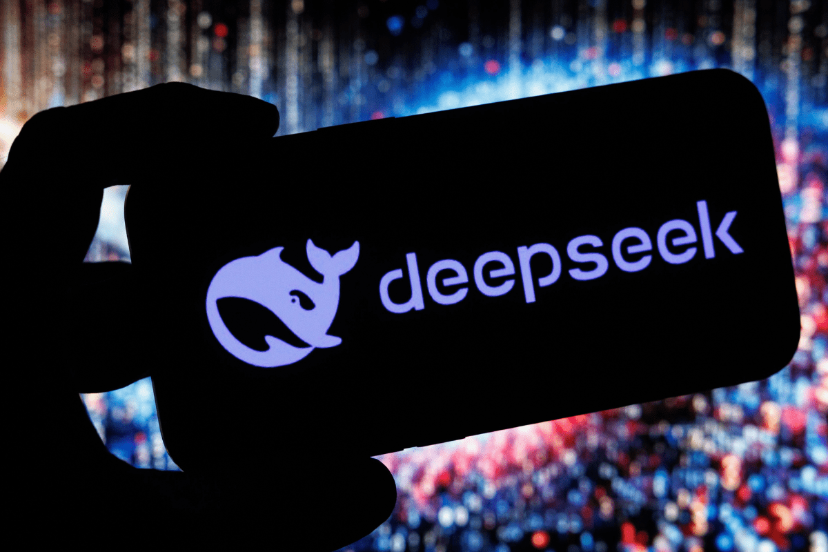 Imagem de uma tela de celular com o logo do DeepSeek iluminado, sendo segurada por uma mão, com fundo de luzes difusas em tons de azul e vermelho.