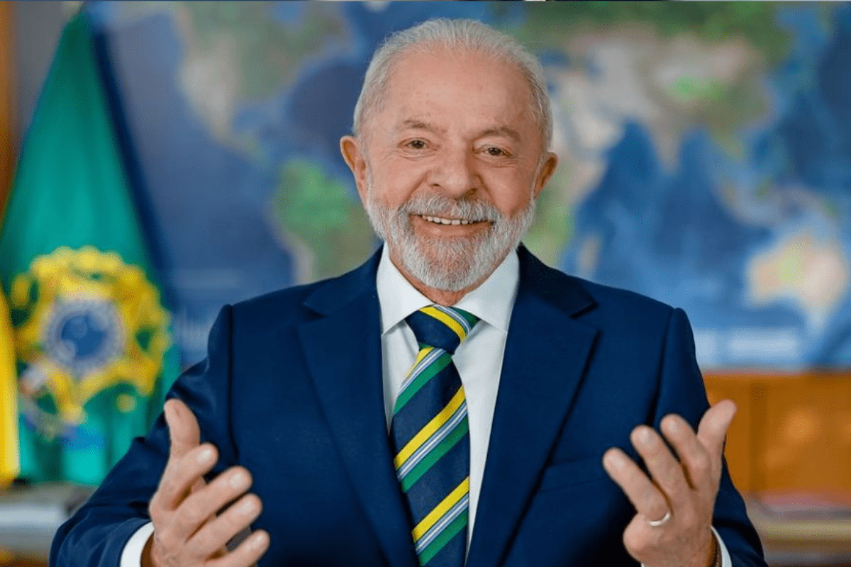 Imagem de um homem idoso sorridente, usando terno azul e gravata listrada, em um ambiente com bandeira do Brasil ao fundo, simbolizando liderança e governança.