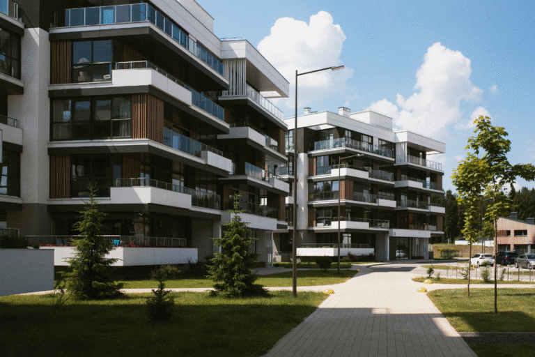Imagem de um conjunto de prédios residenciais modernos com várias unidades, lojas de vidro e varandas amplas, localizado em uma área com árvores e iluminação moderna.