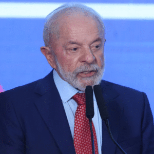 Lula de terno escuro e gravata vermelha fala ao microfone contra um fundo azul e vermelho, parecendo pensativo e engajado.