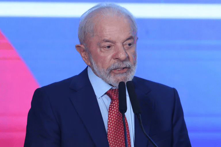 Lula de terno escuro e gravata vermelha fala ao microfone contra um fundo azul e vermelho, parecendo pensativo e engajado.