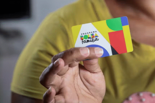 Uma pessoa de camisa amarela segura um cartão colorido com a inscrição "Programa Bolsa Família". O cartão apresenta formas abstratas em verde, vermelho, azul e amarelo.