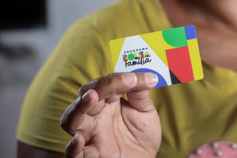 Uma pessoa de camisa amarela segura um cartão colorido com a inscrição "Programa Bolsa Família". O cartão apresenta formas abstratas em verde, vermelho, azul e amarelo.