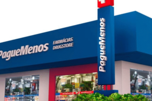 Imagem da fachada de uma farmácia Pague Menos com sua sinalização visível e o interior mostrado pelas janelas, destaque para a loja de produtos médicos e medicamentos.
