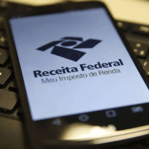 Imagem de um celular sobre um teclado de computador exibindo o logo da Receita Federal e o slogan Meu Imposto de Renda, destacando o tema de declaração de imposto de renda no Brasil.