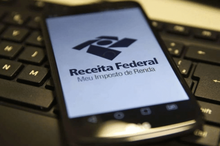 Imagem de um celular sobre um teclado de computador exibindo o logo da Receita Federal e o slogan Meu Imposto de Renda, destacando o tema de declaração de imposto de renda no Brasil.