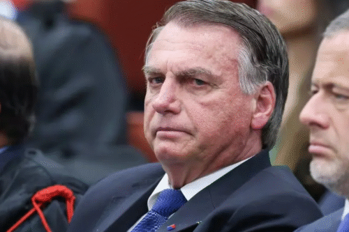 Bolsonaro, com expressão séria, usando terno e gravata, em um ambiente formal, ao lado de outras pessoas.