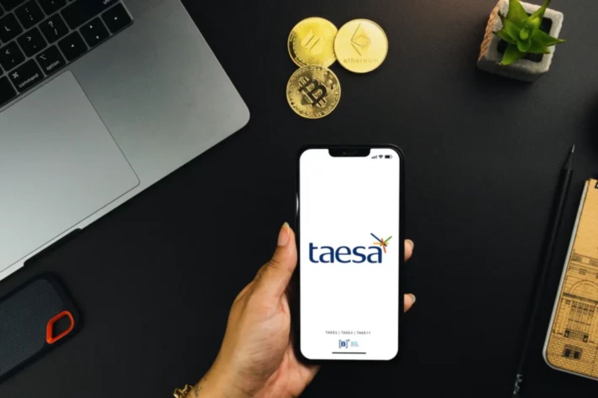 Pessoa segurando um smartphone com a tela exibindo o aplicativo Taesa, ao lado de moedas de bitcoin e altcoins, em um ambiente de escritório com laptop e plantas.