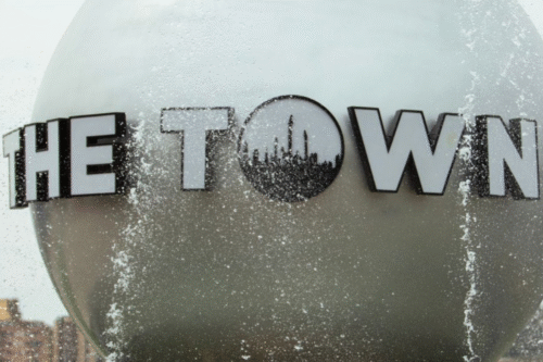 Logotipo de 'The Town' no lado de um prédio, com gotas de água e reflexo no vidro, representando um ponto turístico ou local conhecido, sob um céu nublado.