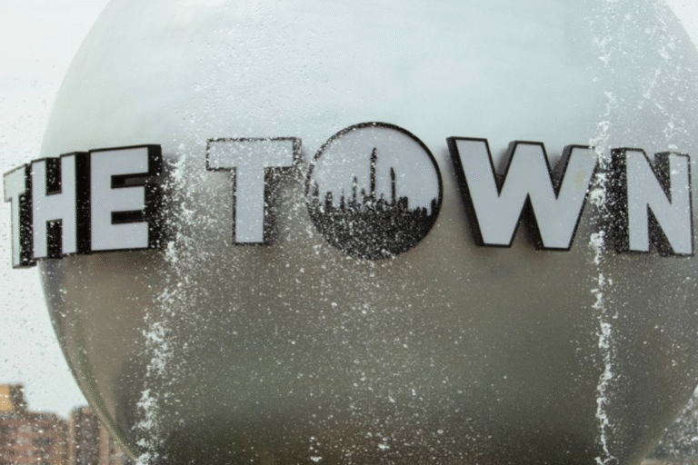 Logotipo de 'The Town' no lado de um prédio, com gotas de água e reflexo no vidro, representando um ponto turístico ou local conhecido, sob um céu nublado.