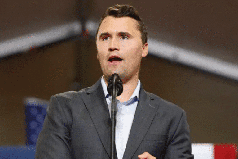 Charlie Kirk de terno e gravata falando em um microfone durante um evento ou apresentação.