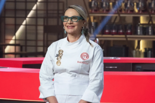 Participante Daniela sorrindo em programa de culinária, vestida com jaleco branco e óculos, em ambiente de cozinha com bancada vermelha ao fundo.