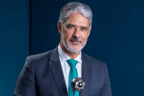William Bonner de terno cinza e gravata azul, com cabelo grisalho e barba, sorrindo levemente enquanto fala na câmera durante evento formal
