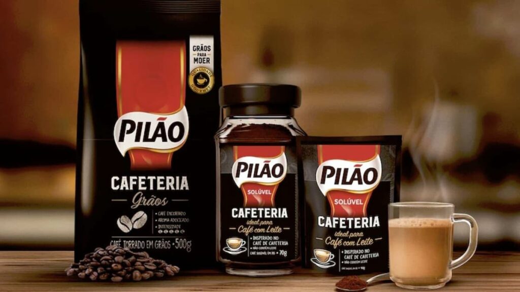 Pacotes de café Pilão Cafeteria em grãos e solúvel, ao lado de xícara de café com leite fumegante.