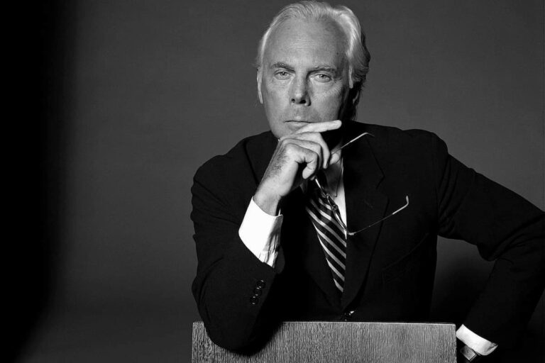 Retrato em preto e branco de Giorgio Armani em terno, mão no queixo segurando óculos, fundo escuro.