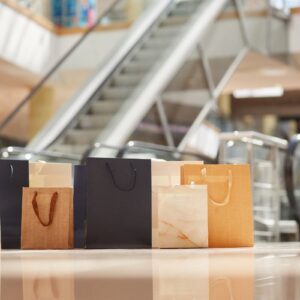 Sacolas de compras elegantes, com escadas e ambiente de shopping ao fundo.