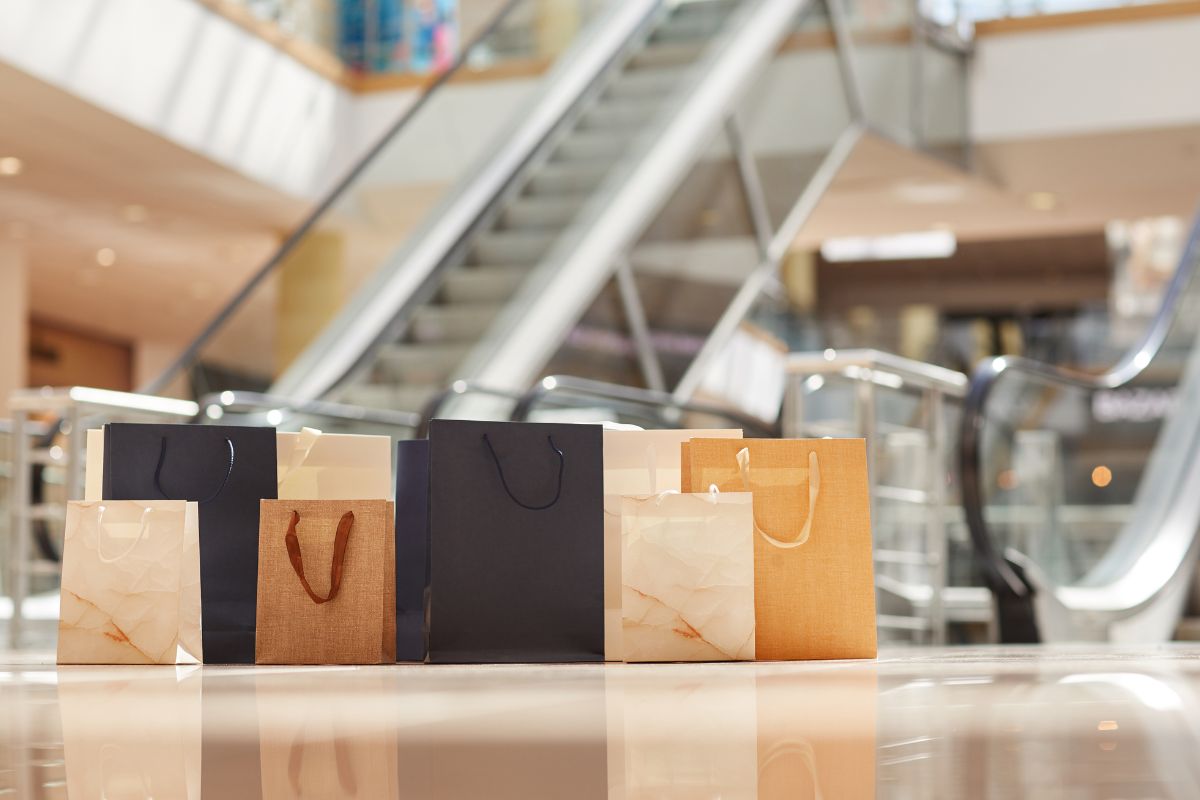 Sacolas de compras elegantes, com escadas e ambiente de shopping ao fundo.