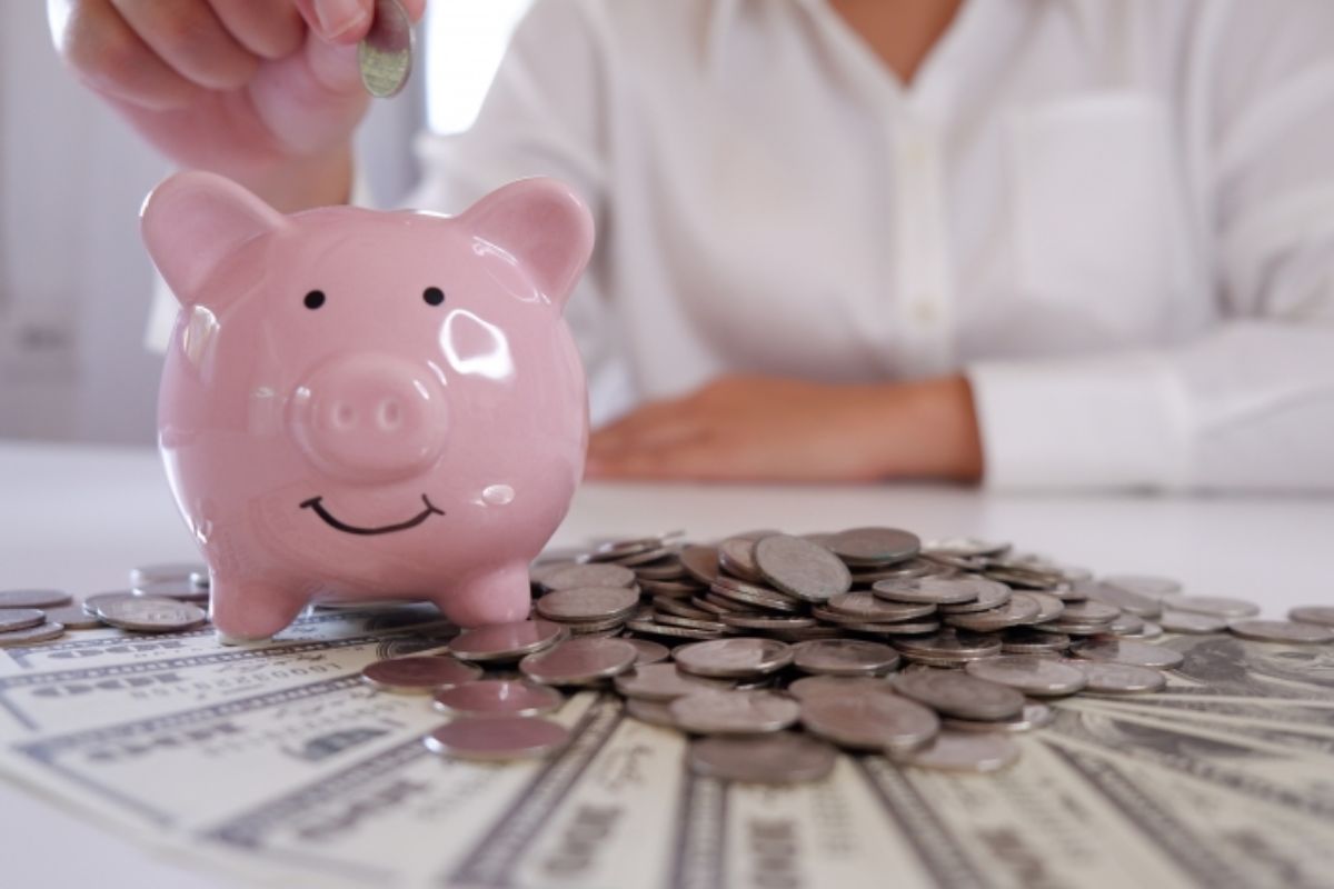 Pessoa colocando uma moeda no cofrinho de porquinho rosa com sorriso, cercado de dinheiro em papel moeda e moedas de metal, simbolizando economia e finanças pessoais.
