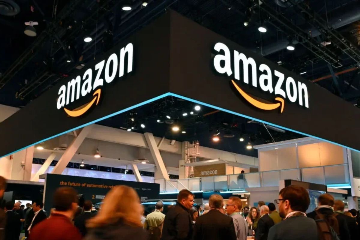 Imagem do estande da Amazon na feira de tecnologia com diversas pessoas ao redor, destaque para o logo da Amazon no teto, em ambiente de evento corporativo.