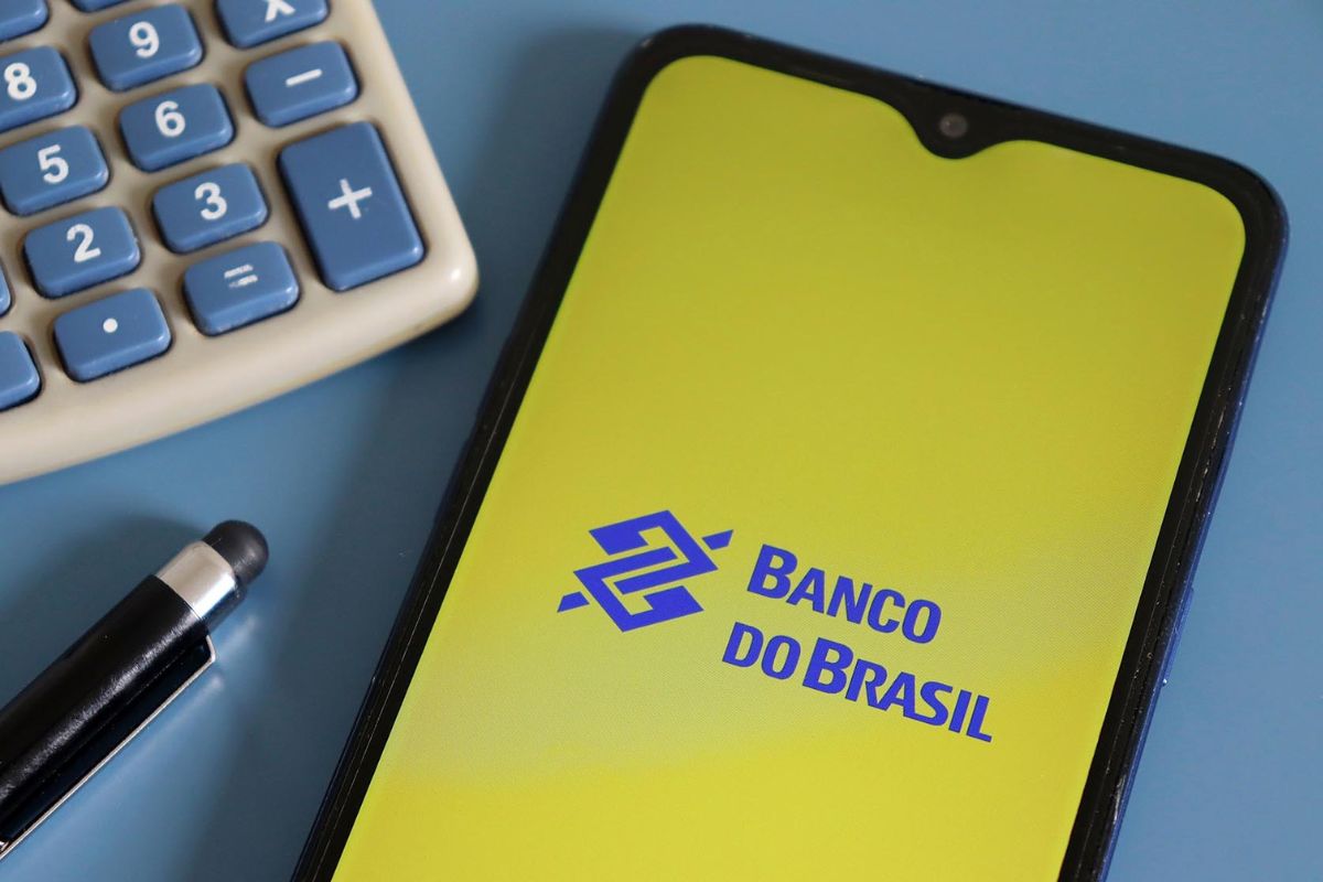 Celular exibindo o aplicativo do Banco do Brasil na tela, ao lado de uma calculadora e uma caneta em uma mesa azul, simbolizando serviços bancários digitais.