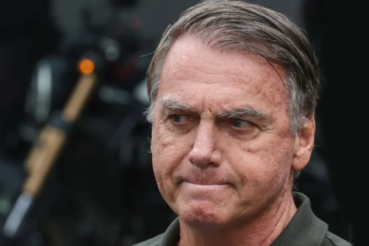 Imagem de Bolsonaro com expressão séria, cabelo grisalho penteado para trás, pele com sinais de envelhecimento e um fundo escuro desfocado.