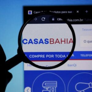 Imagem de uma pessoa segurando uma lupa que destaca o logo do site Casas Bahia na tela de um computador, promovendo compras online de móveis, eletrônicos e eletrodomésticos.