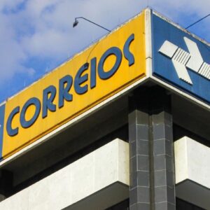 Imagem da fachada de uma agência dos Correios com identidade visual amarela, azul e o logo característico em destaque, localizado em um edifício alto com céu azul ao fundo.