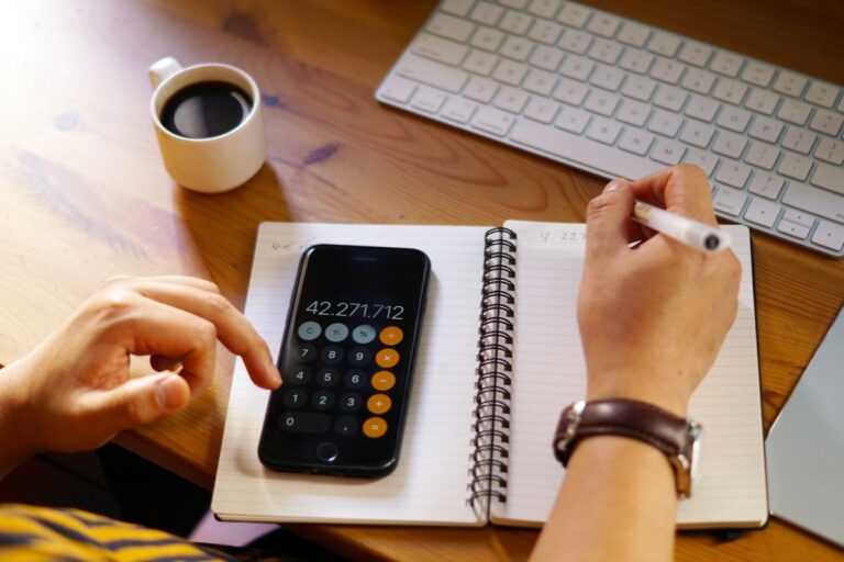 Pessoa usando uma calculadora financeira ao lado de um computador, caderno e uma xícara de café na bancada de madeira, ambiente de escritório.