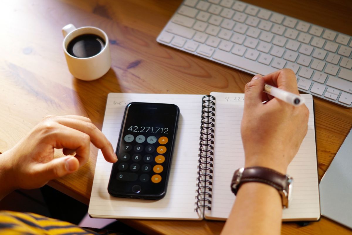 Pessoa usando uma calculadora financeira ao lado de um computador, caderno e uma xícara de café na bancada de madeira, ambiente de escritório.