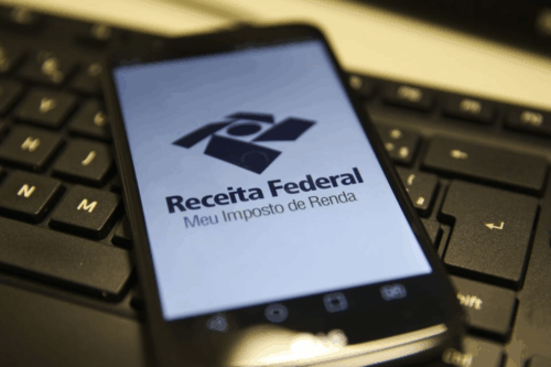 Imagem de um smartphone com a tela exibindo o logo e a mensagem da Receita Federal, sobre Meu Imposto de Renda, colocado sobre um teclado de computador.