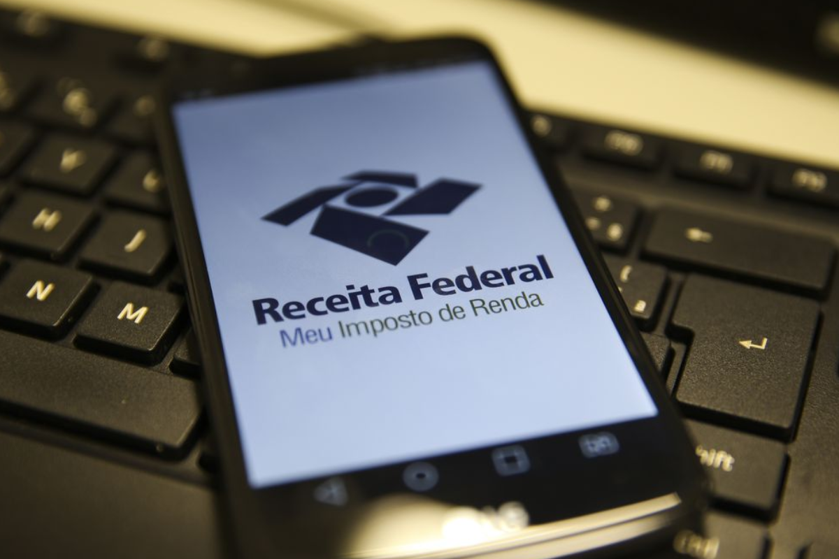 Imagem de um smartphone com a tela exibindo o logo e a mensagem da Receita Federal, sobre Meu Imposto de Renda, colocado sobre um teclado de computador.