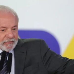 Lula de cabelos brancos e barba, vestindo terno escuro, fazendo uma apresentação ou discurso em um evento, com fundo colorido e iluminado.